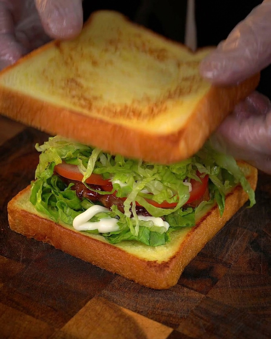 Gloved hands gently placing a golden-brown slice of toasted bread on top of a sandwich filled with fresh lettuce, tomato, mayonnaise, and glazed chicken.