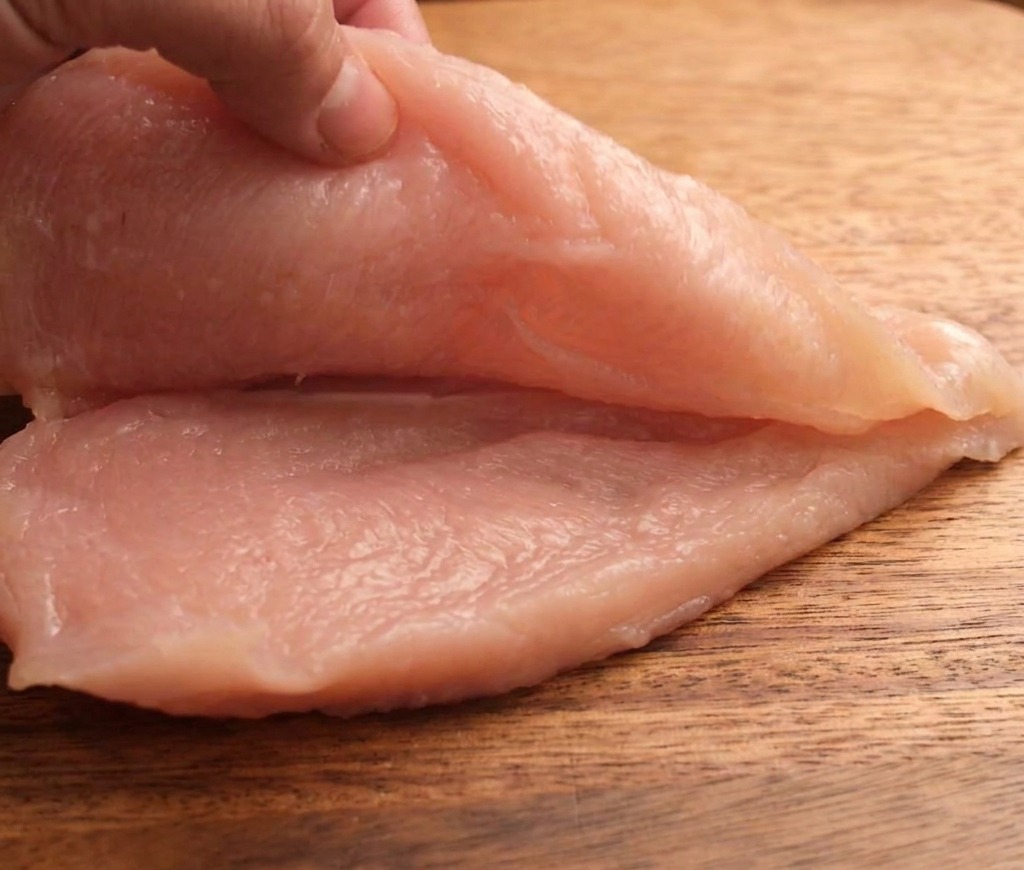 A hand slicing a raw chicken breast horizontally into two thinner pieces on a wooden cutting board.