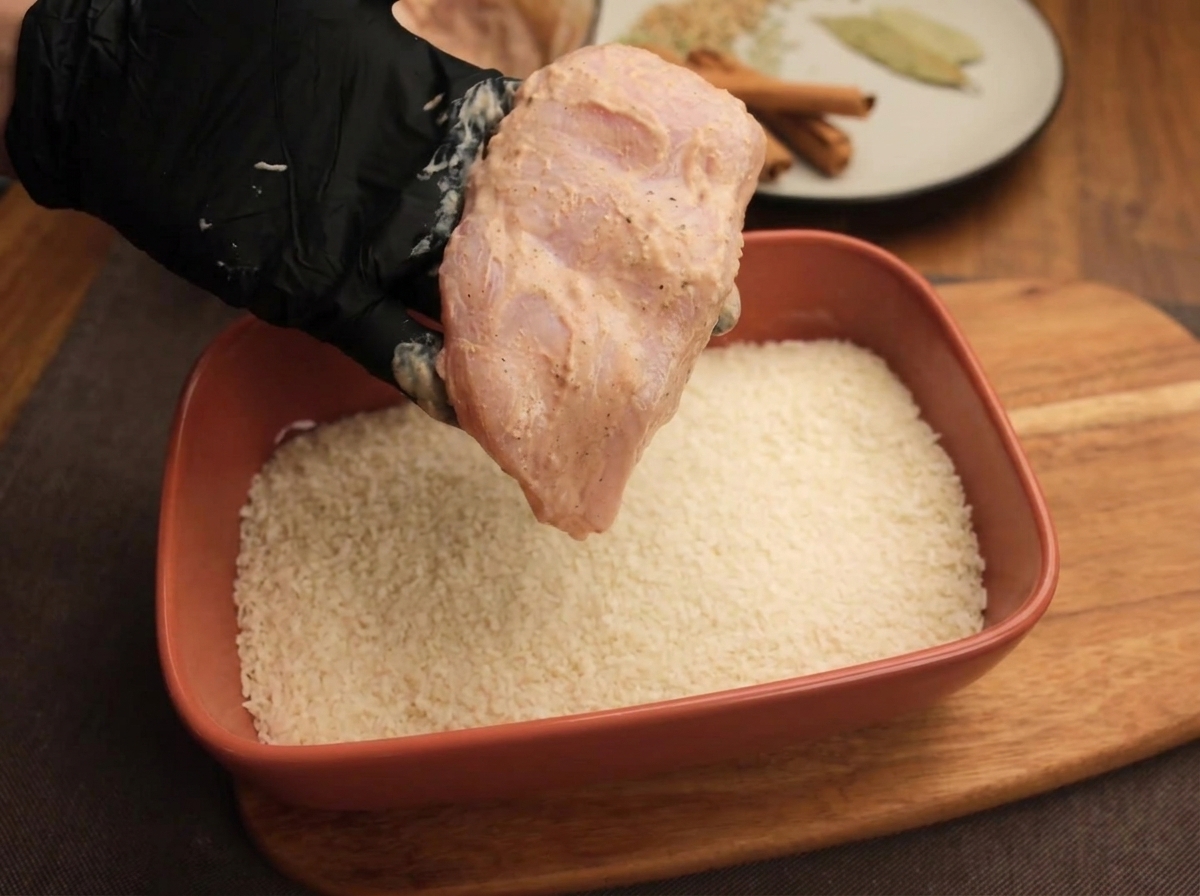 A gloved hand holding a yogurt-marinated chicken cutlet over a rectangular tray filled with white breadcrumbs.