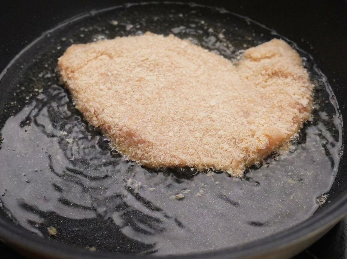 A breaded chicken breast piece bubbling in hot frying oil inside a black skillet.