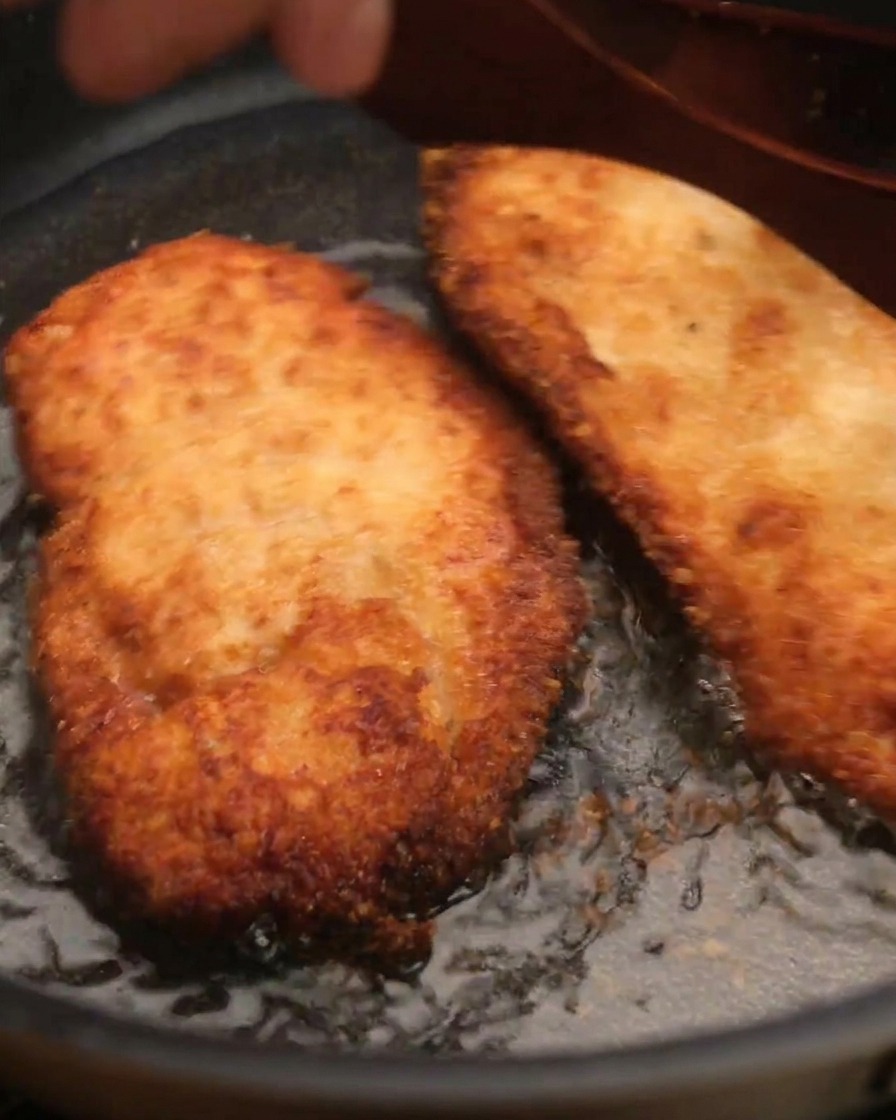A piece of crispy fried chicken being lifted from a pan of oil using a wooden spatula.