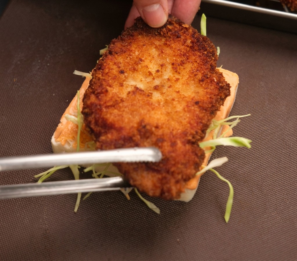 A hand and metal tongs placing a crispy fried chicken cutlet onto a slice of white bread topped with orange sauce and thin cabbage strips.