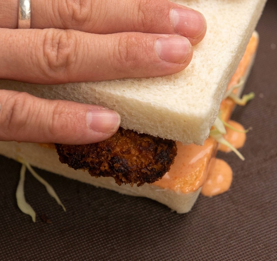 Hands pressing down on the top slice of a fried chicken sandwich, with cabbage and orange spicy mayo visible between the bread slices.