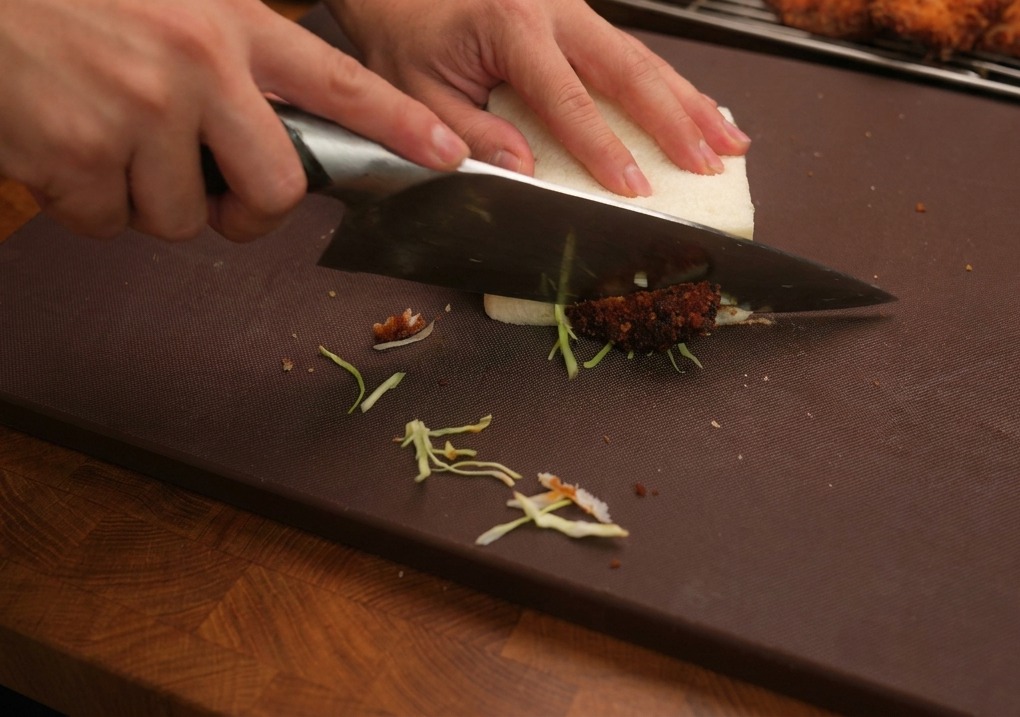 A large kitchen knife slicing through a thick fried chicken sandwich on a dark brown cutting board, with extra fried chicken pieces in the background.