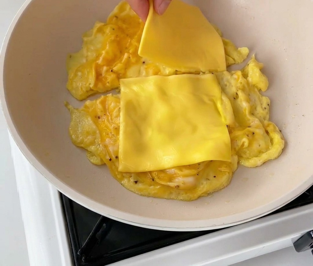 A hand placing two slices of yellow cheddar cheese onto portions of soft scrambled eggs in a light-colored frying pan.