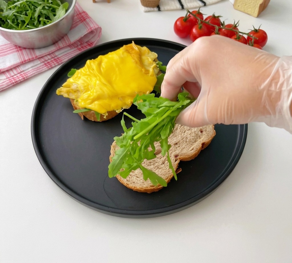 A hand placing fresh green arugula leaves onto a slice of toasted multigrain bread on a matte black plate.
