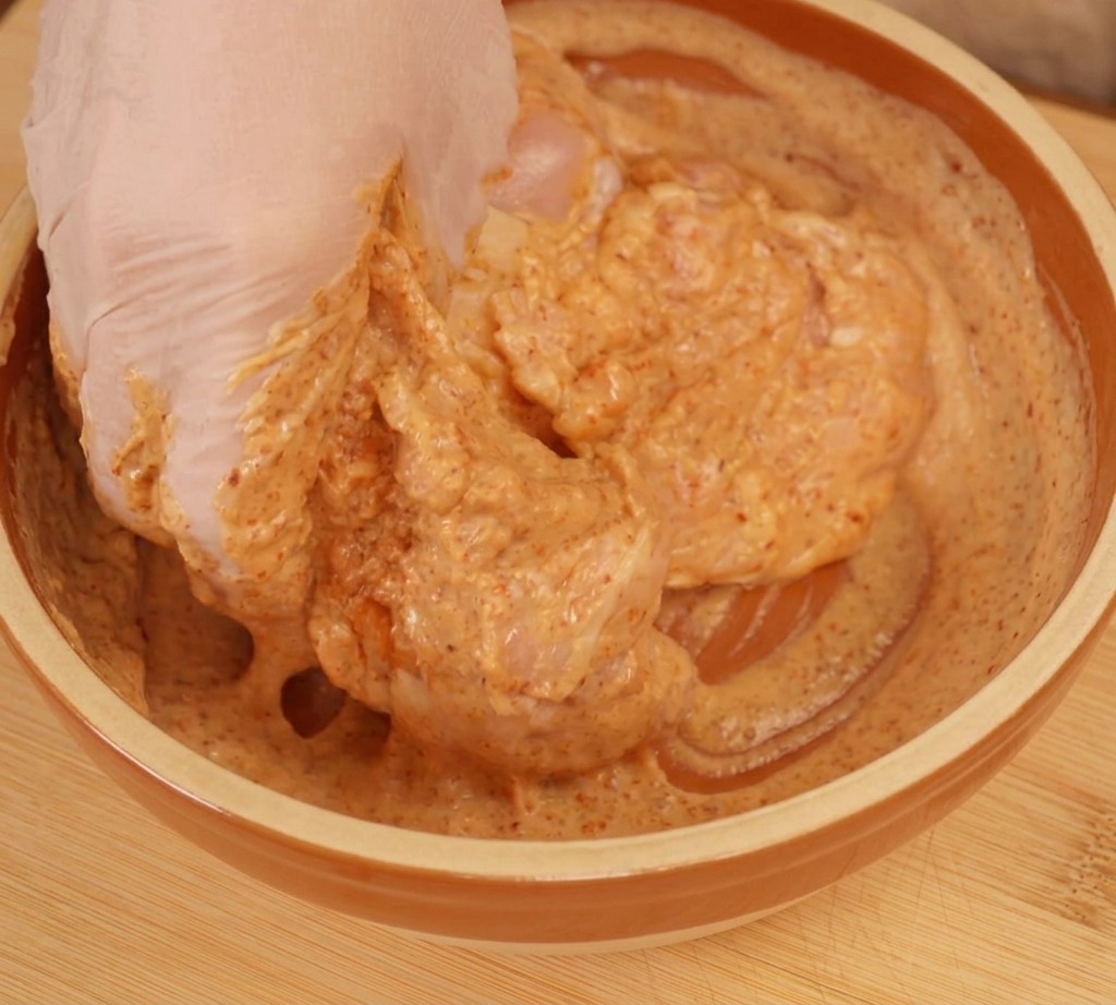 A gloved hand mixing raw chicken pieces thoroughly in a creamy, orange-colored yogurt marinade.