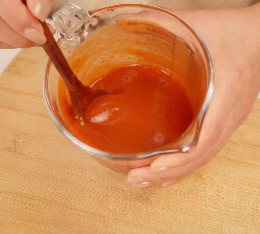 Hands using a wooden spoon to stir a thick, bright red sauce mixture in a glass measuring cup.