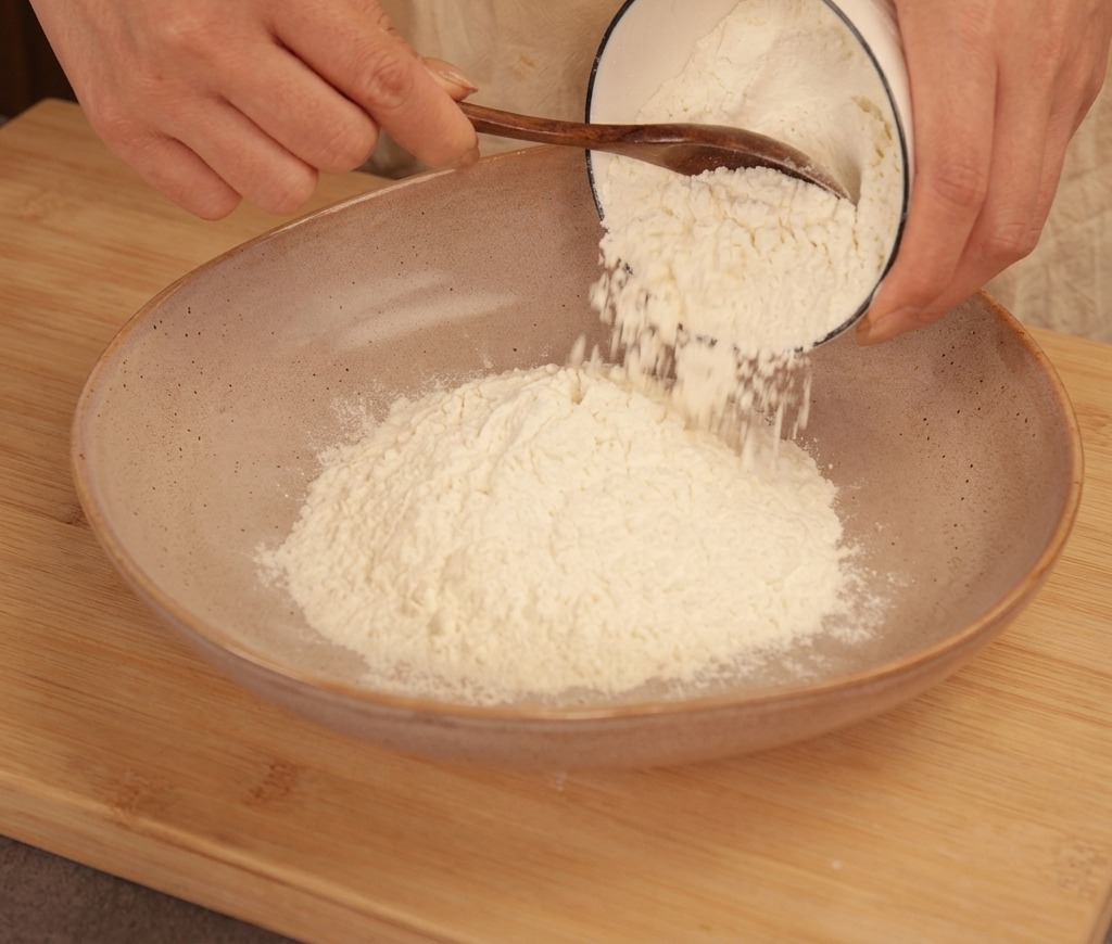 A person pouring white cornstarch from a small bowl into a larger ceramic bowl filled with all-purpose flour.