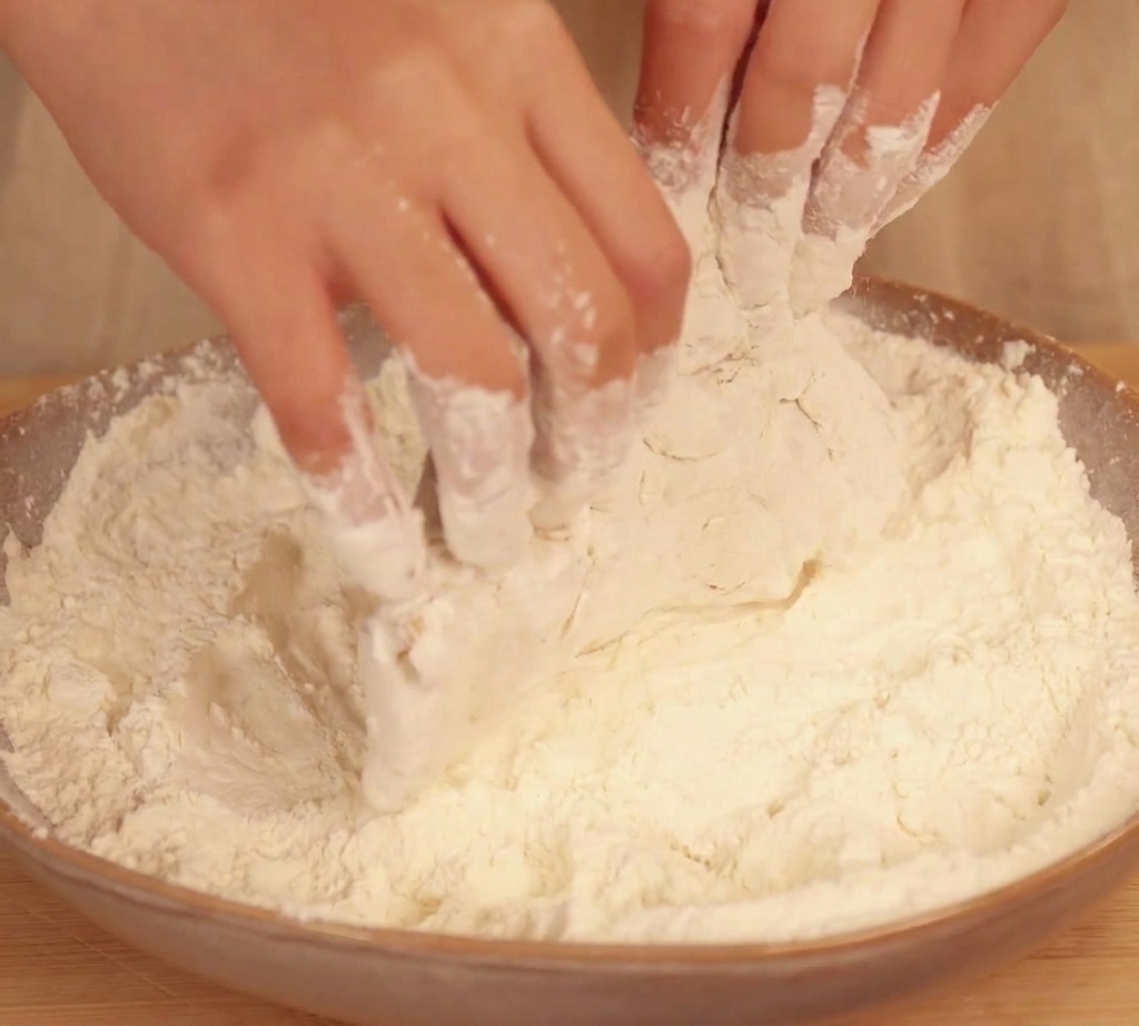 Hands tossing and pressing a wet piece of chicken into a bowl of dry flour to create a thick, flaky breading.