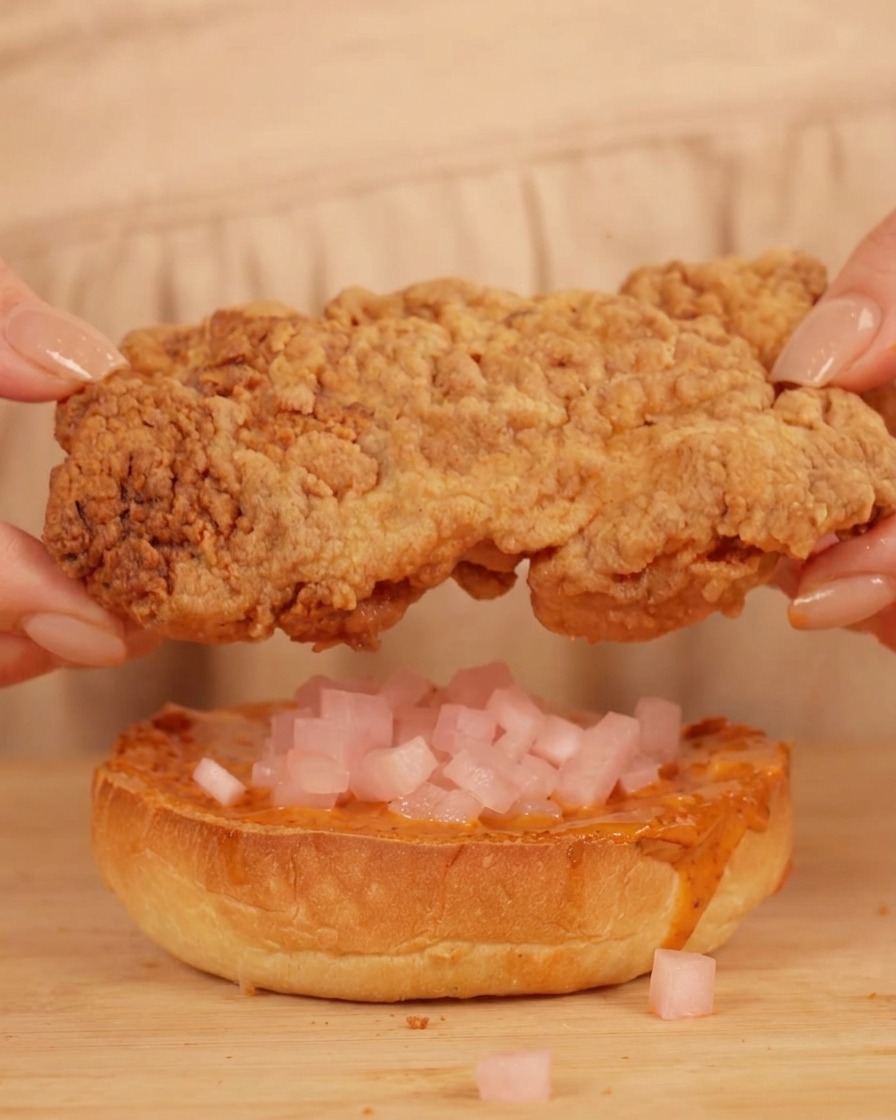 A pair of hands holding a large, golden-brown fried chicken fillet directly above a toasted burger bun topped with sauce and small pink pickled radish cubes.