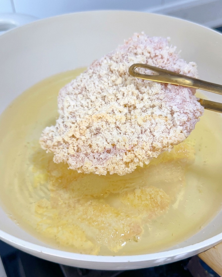 A frozen breaded chicken cutlet being lowered into bubbling hot oil using gold tongs.