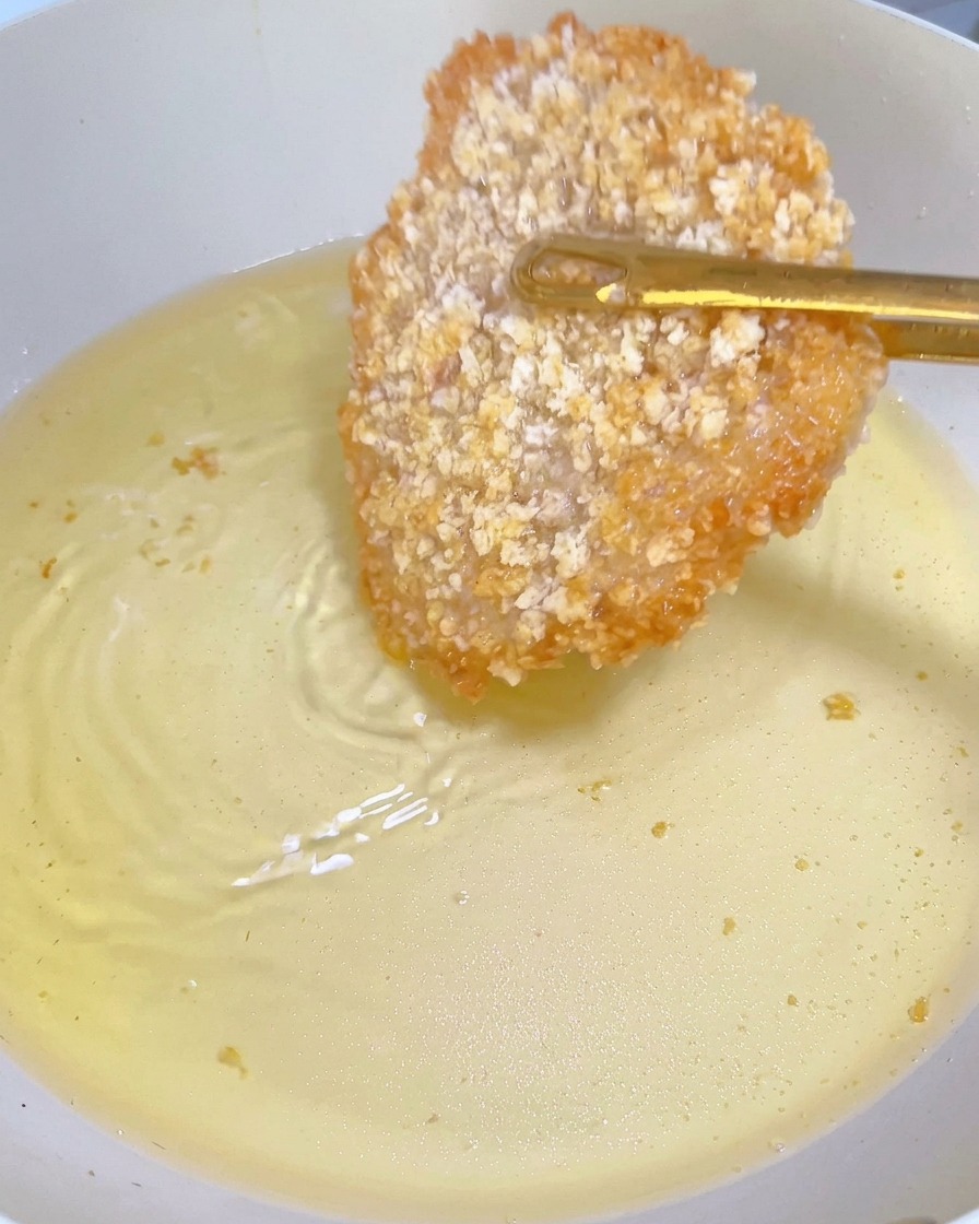 A partially fried chicken cutlet showing a golden-brown crust, being flipped with gold tongs in a pan of hot oil.