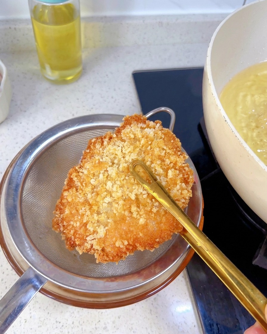A fully fried, golden-brown chicken cutlet held with tongs over a metal strainer to drain excess oil.