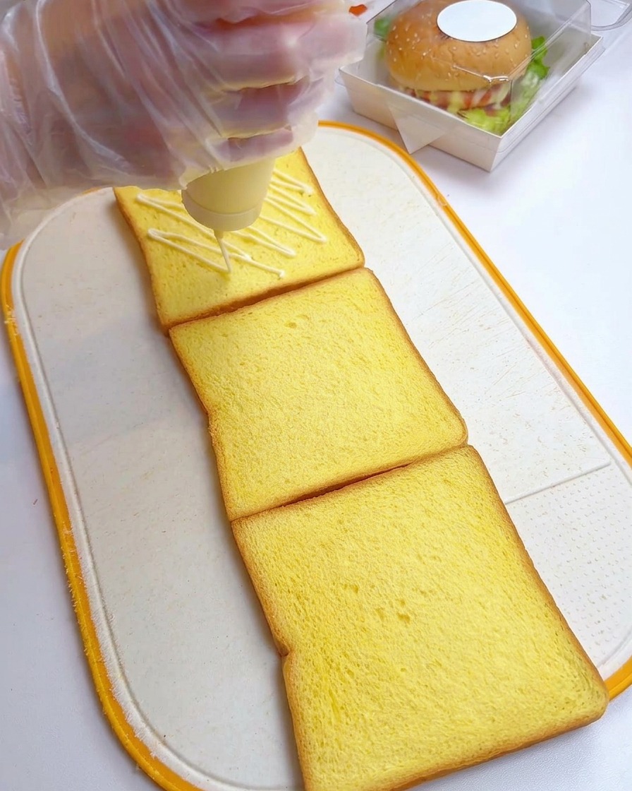 A gloved hand squeezing mayonnaise in a zigzag pattern onto three slices of yellow toast on a cutting board.