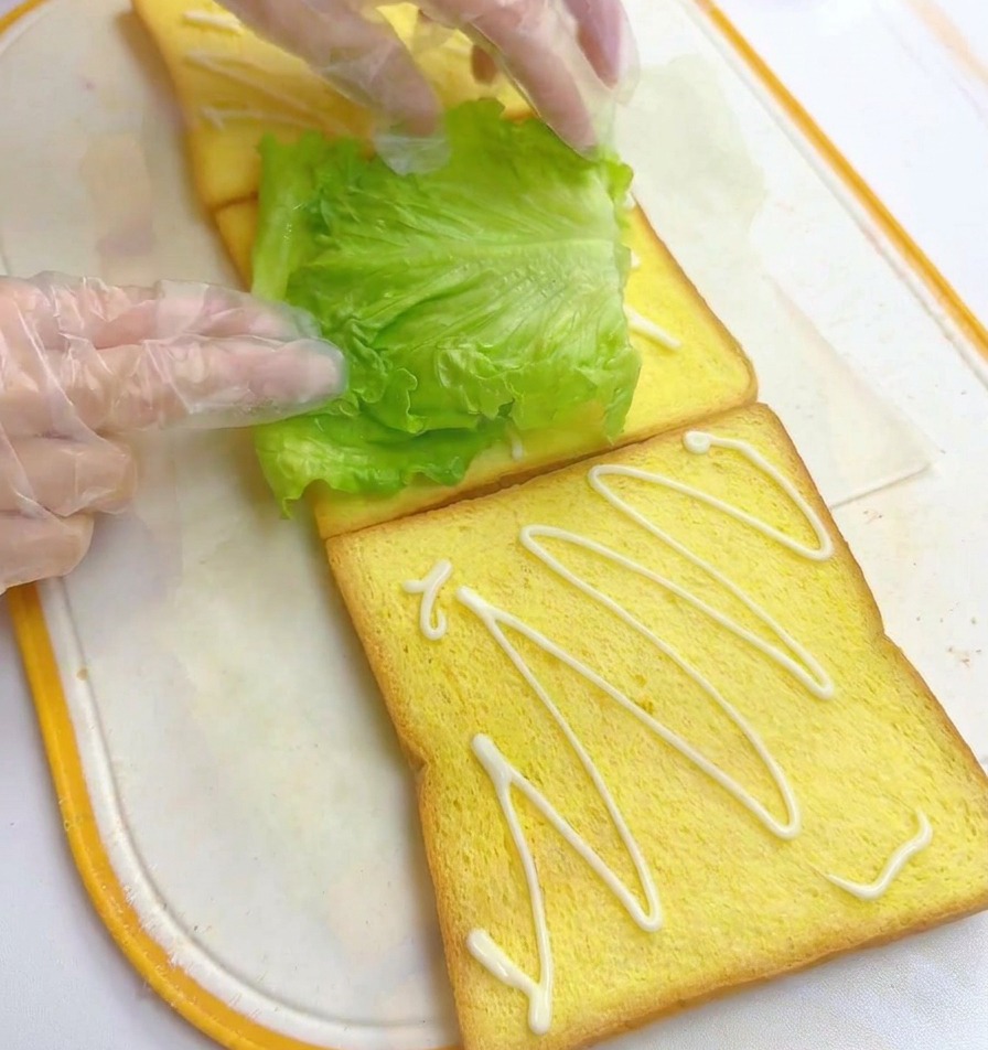 Gloved hands placing a fresh green lettuce leaf on a slice of yellow toast, with another slice of toast drizzled with mayonnaise next to it.