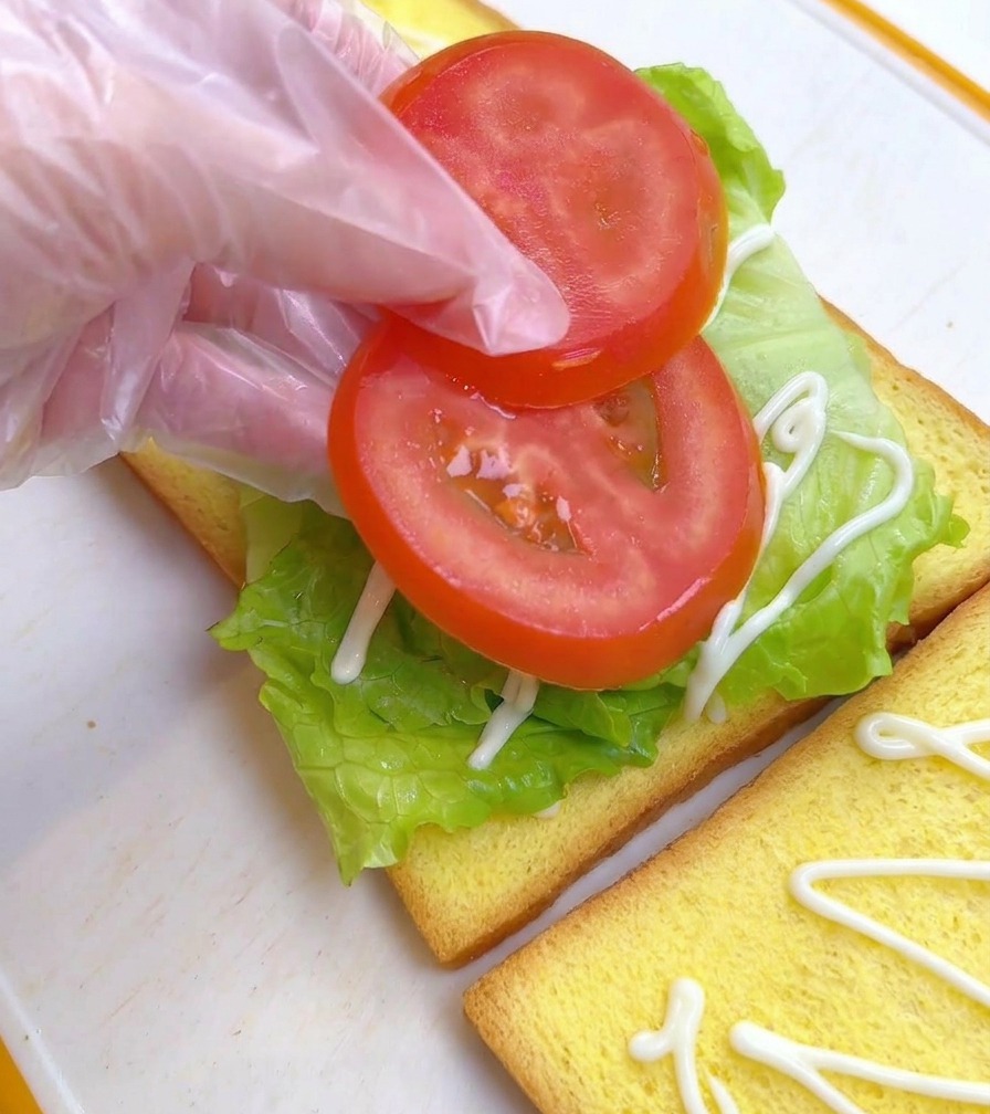 Gloved fingers gently placing two round, red tomato slices on top of a lettuce leaf on a piece of yellow toast.