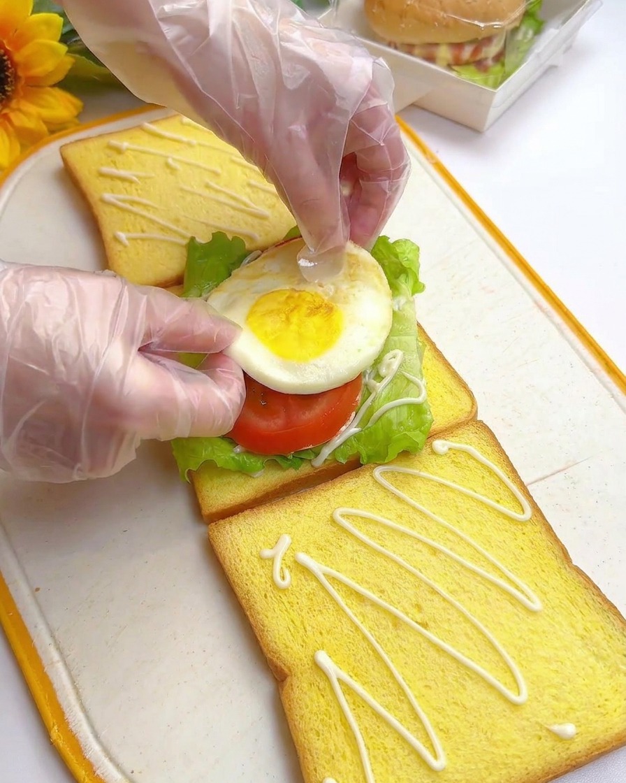 Gloved hands carefully placing a perfectly fried egg on top of fresh tomato slices and green lettuce on a piece of yellow toast.
