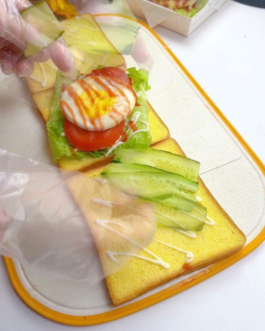 Gloved hands arranging thin diagonal slices of fresh green cucumber onto a slice of yellow toast.