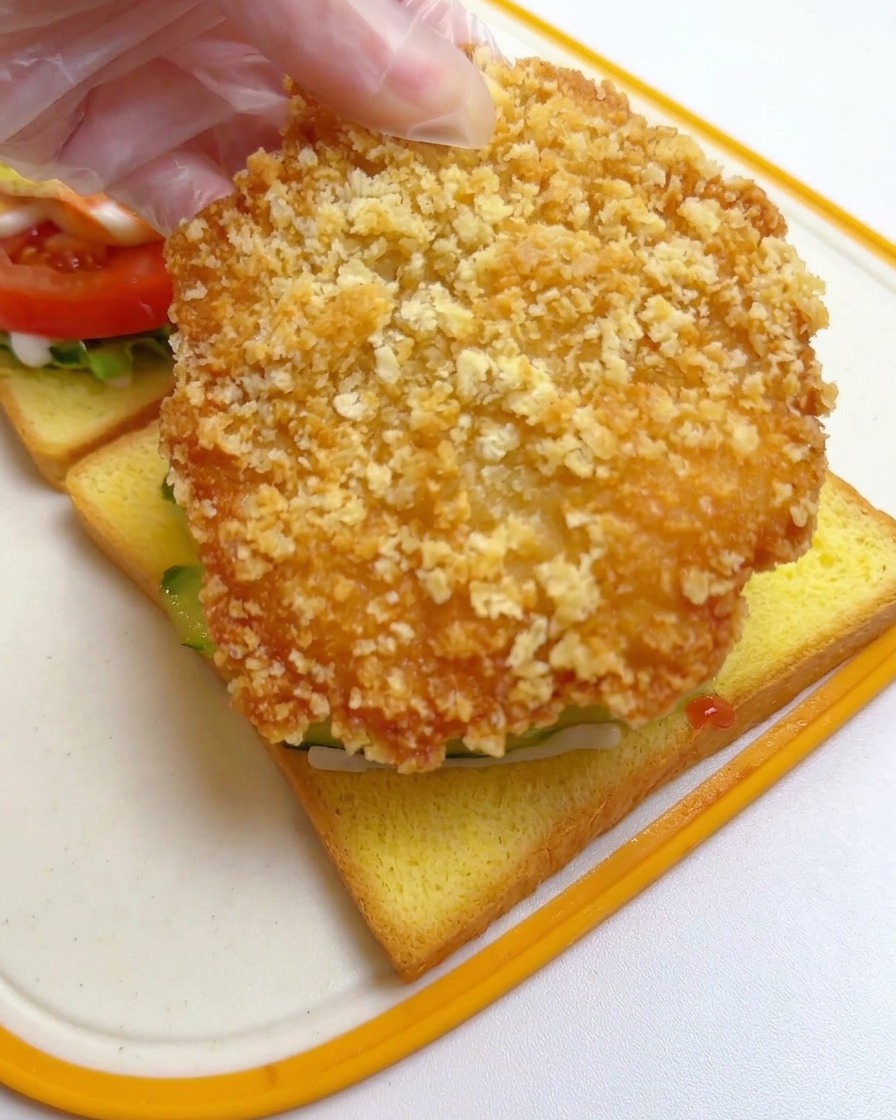 A hand wearing a clear plastic glove placing a large, golden-brown crispy fried chicken cutlet on top of cucumber slices on yellow toast.