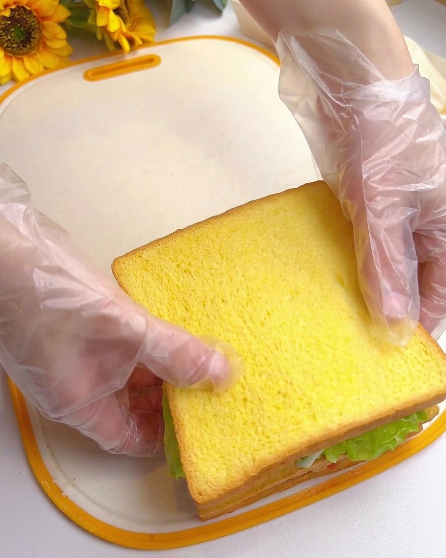 Two gloved hands pressing down on the top slice of yellow toast, completing a tall, stacked, multi-layered sandwich.