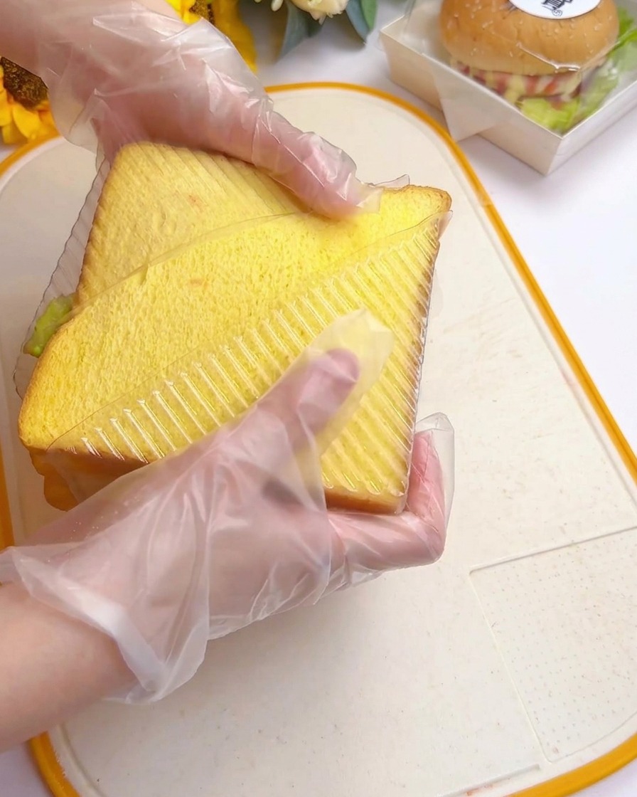 Gloved hands pressing a tall layered sandwich into a clear plastic triangular mold on a cutting board.