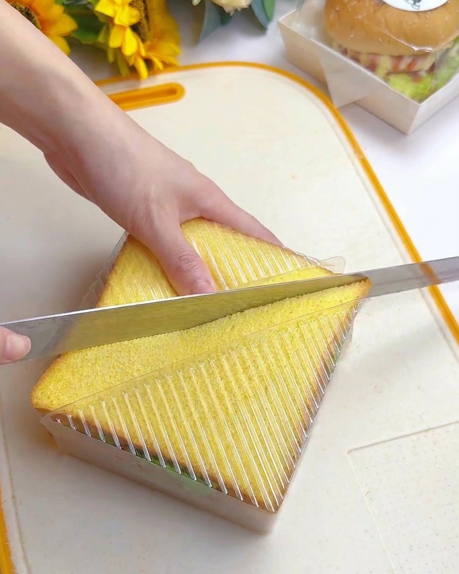 A hand using a long serrated knife to slice diagonally through a tall sandwich encased in a clear plastic guide.