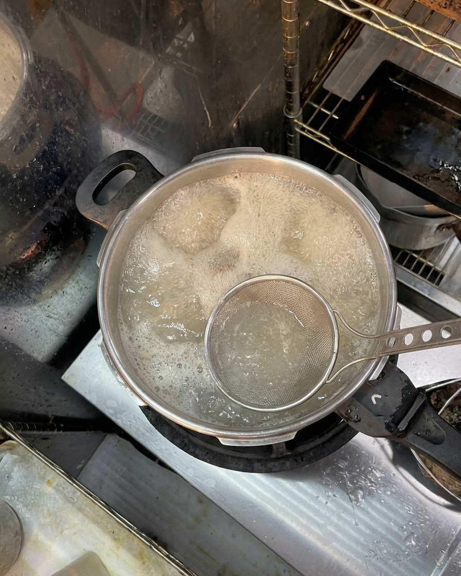 A metal mesh skimmer scooping brownish foam and impurities from the surface of boiling water containing bones.