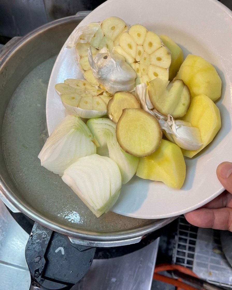 A plate holding halved garlic heads, chopped onions, ginger slices, and potato chunks being held over a pot of boiling broth.