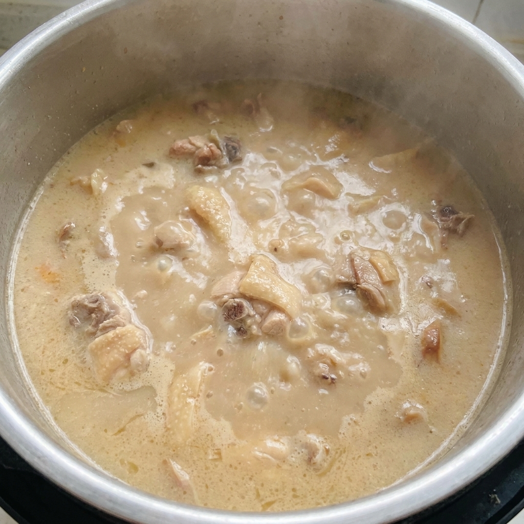 A pot of vigorously boiling, thick, opaque, milky-white tonkotsu broth with meat pieces visible in the bubbling liquid.