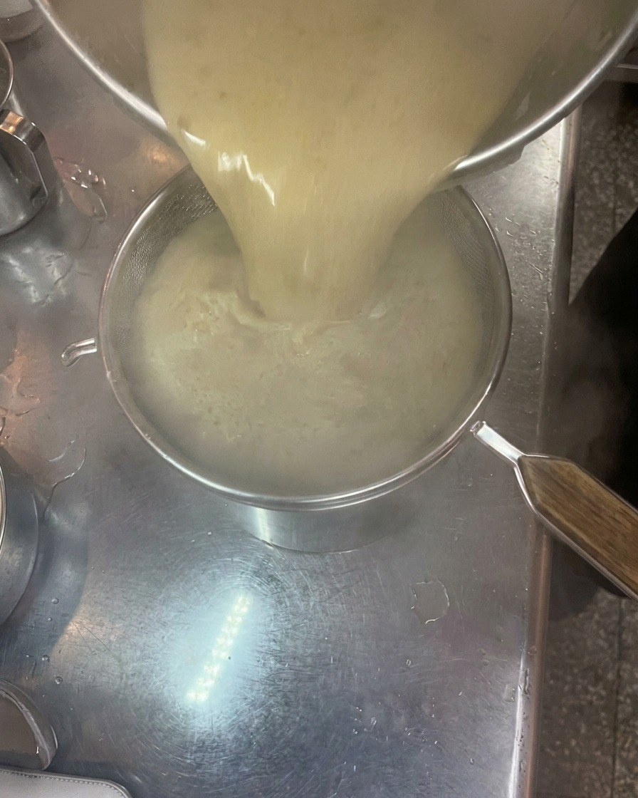 Thick, creamy tonkotsu broth being poured from a pot through a fine mesh strainer into another pot.