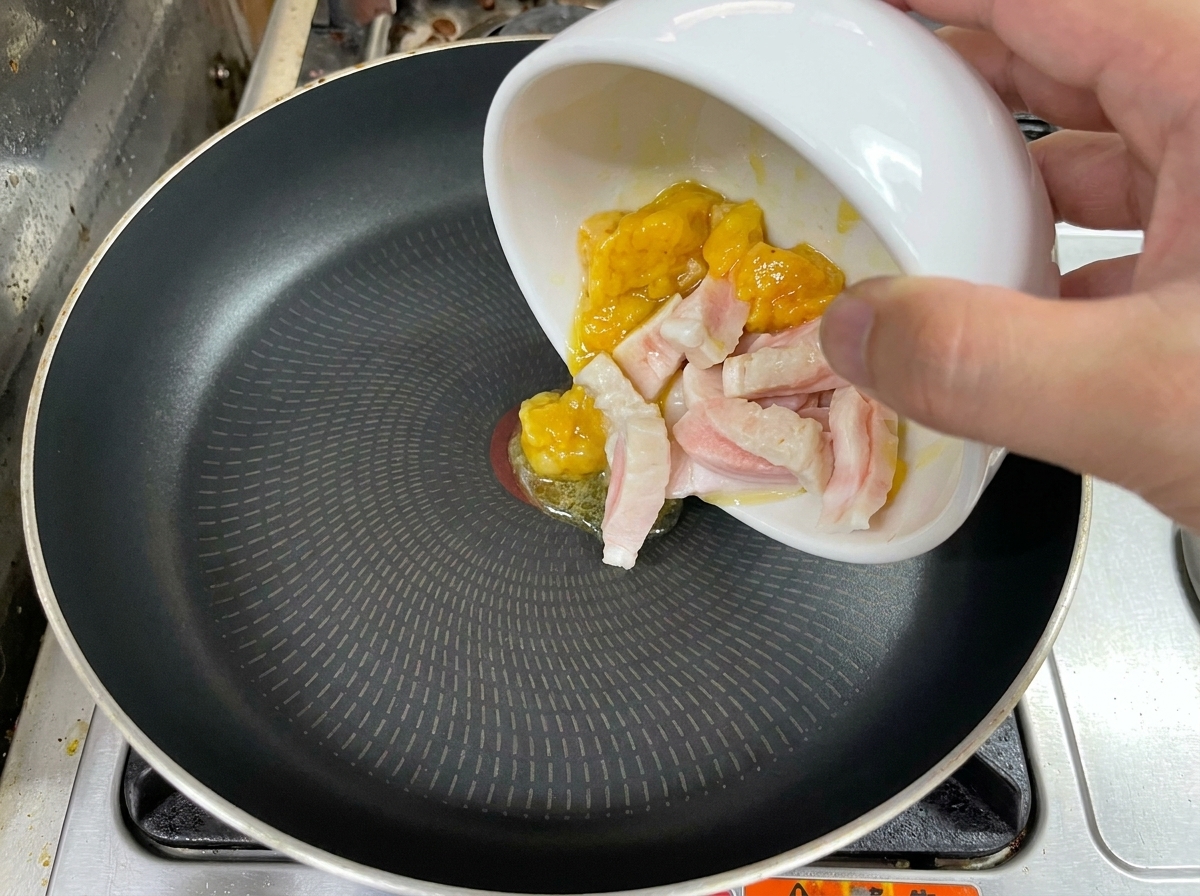 A hand pouring pieces of raw chicken and pork fat from a white bowl into a black frying pan.
