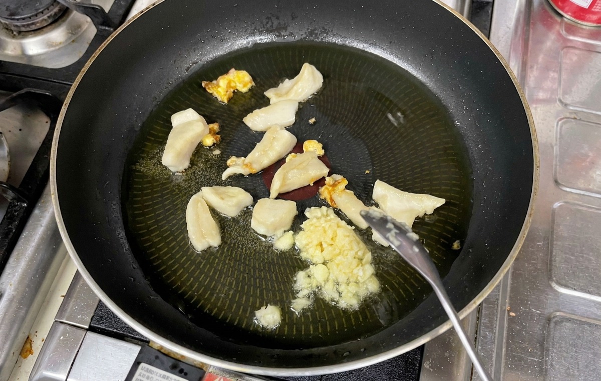 Chopped garlic being stirred with a spoon alongside rendered pieces of fat in a pan of hot oil.