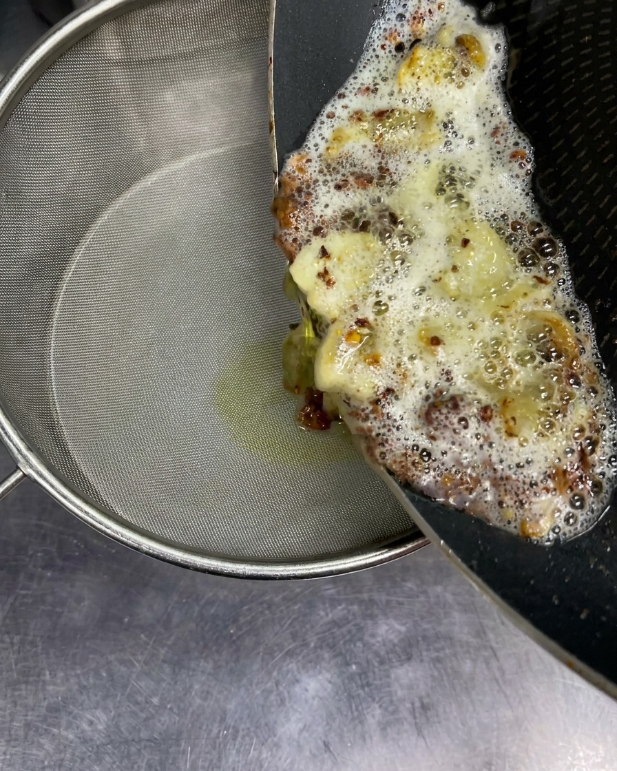 Hot, bubbling aroma oil with fried garlic bits being poured from a pan into a fine mesh sieve.