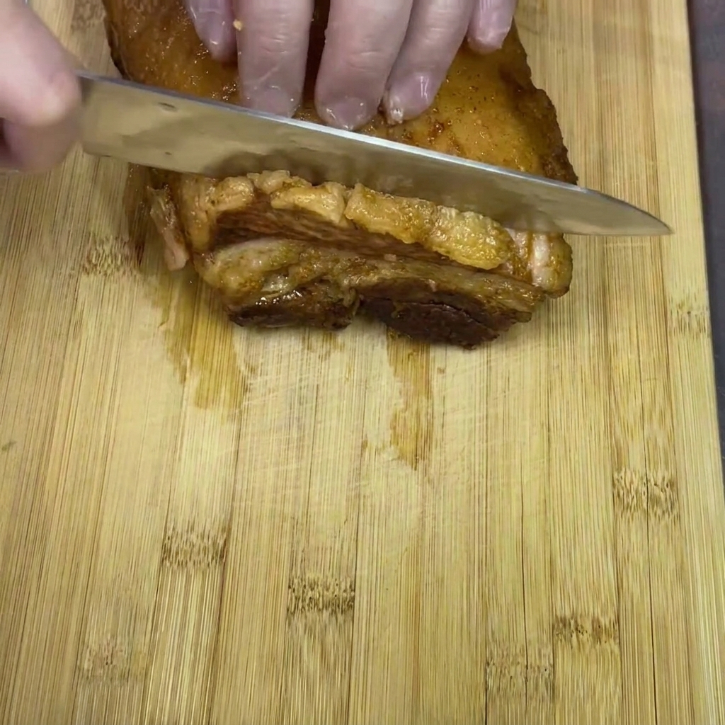 Gloved hands slicing a block of cooked braised pork belly into thick pieces on a wooden cutting board.