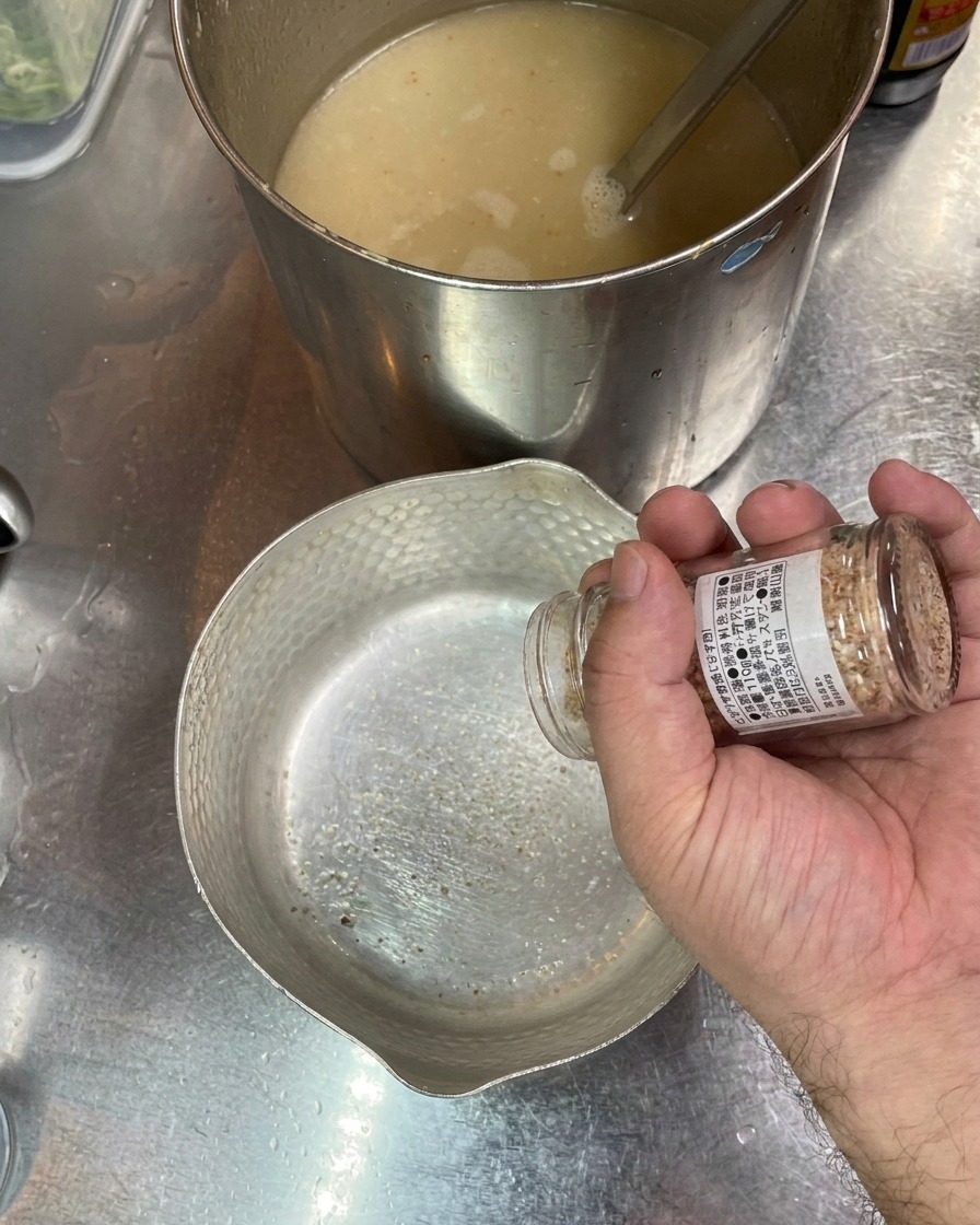 A hand shaking salt from a small glass jar into an empty metal bowl, with a pot of broth in the background.