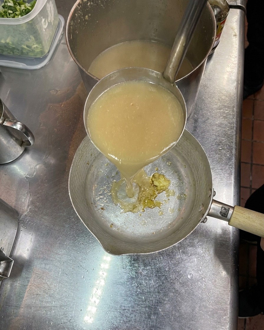 A metal ladle pouring cloudy, rich hot broth into a small saucepan that contains mashed seasonings and garlic.