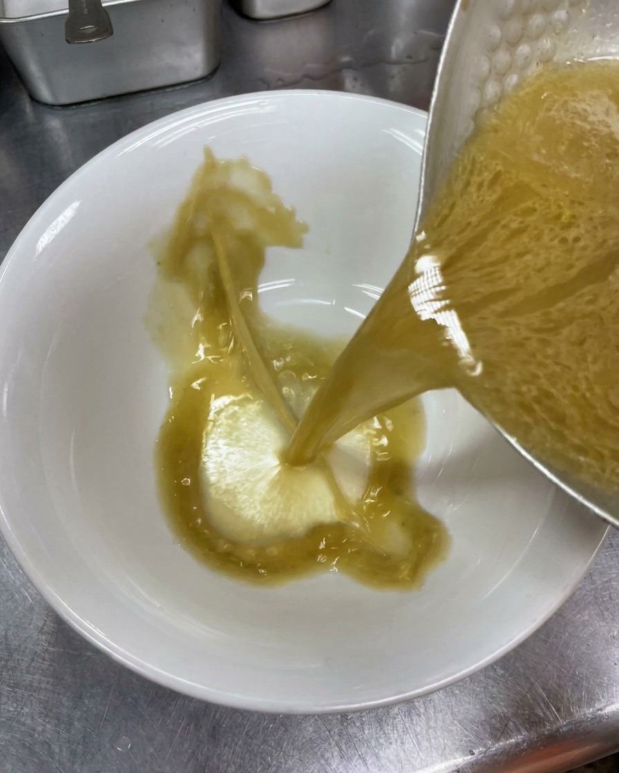 Hot, opaque seasoned tonkotsu broth being poured from a metal pot into a clean white ceramic ramen bowl.