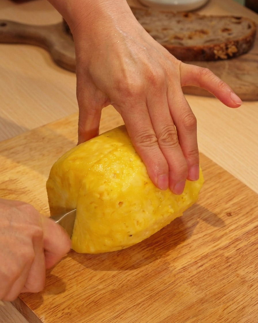 A persons hands using a knife to slice a fresh peeled pineapple on a wooden cutting board.