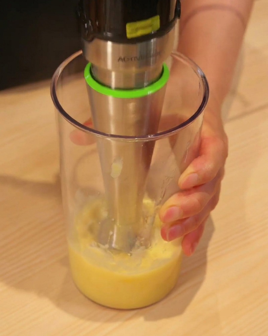 A handheld immersion blender pureeing yellow pineapple chunks into juice in a tall plastic cup.