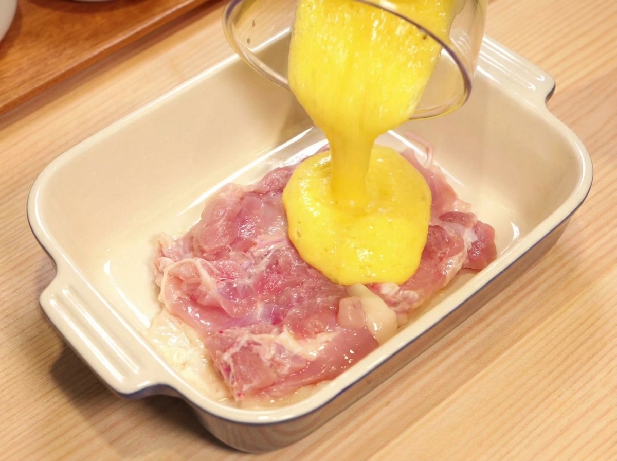 Pouring bright yellow blended pineapple juice from a plastic container over a raw, pink boneless chicken thigh in a white ceramic dish.