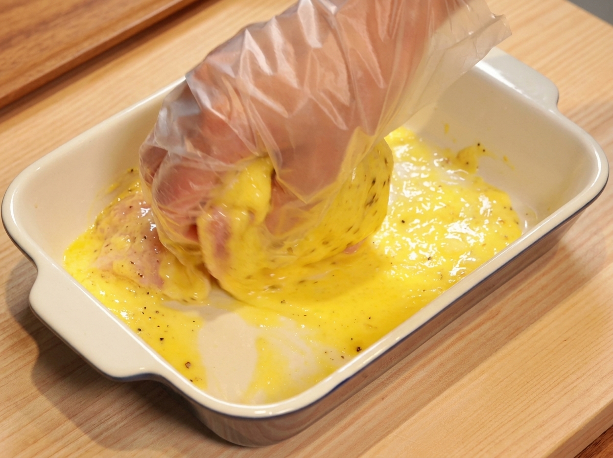A hand wearing a clear plastic glove massaging a raw chicken thigh in a bright yellow pineapple marinade.