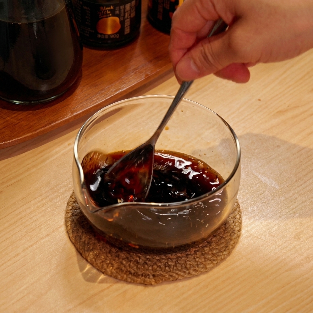 A hand using a metal spoon to stir a dark, mixed liquid sauce in a small glass bowl.