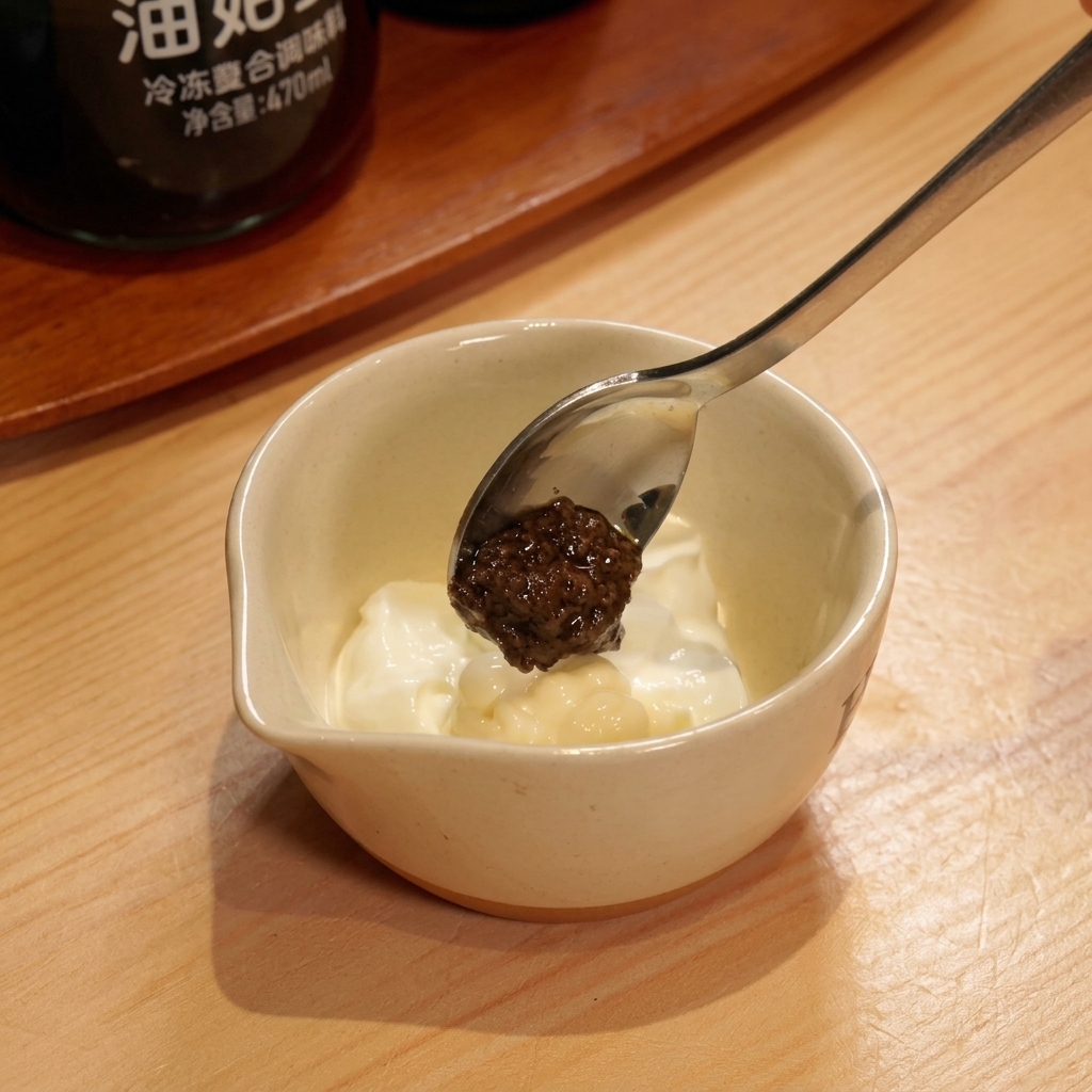 A spoon holding dark black truffle paste hovering over a small bowl of creamy white yogurt dressing.
