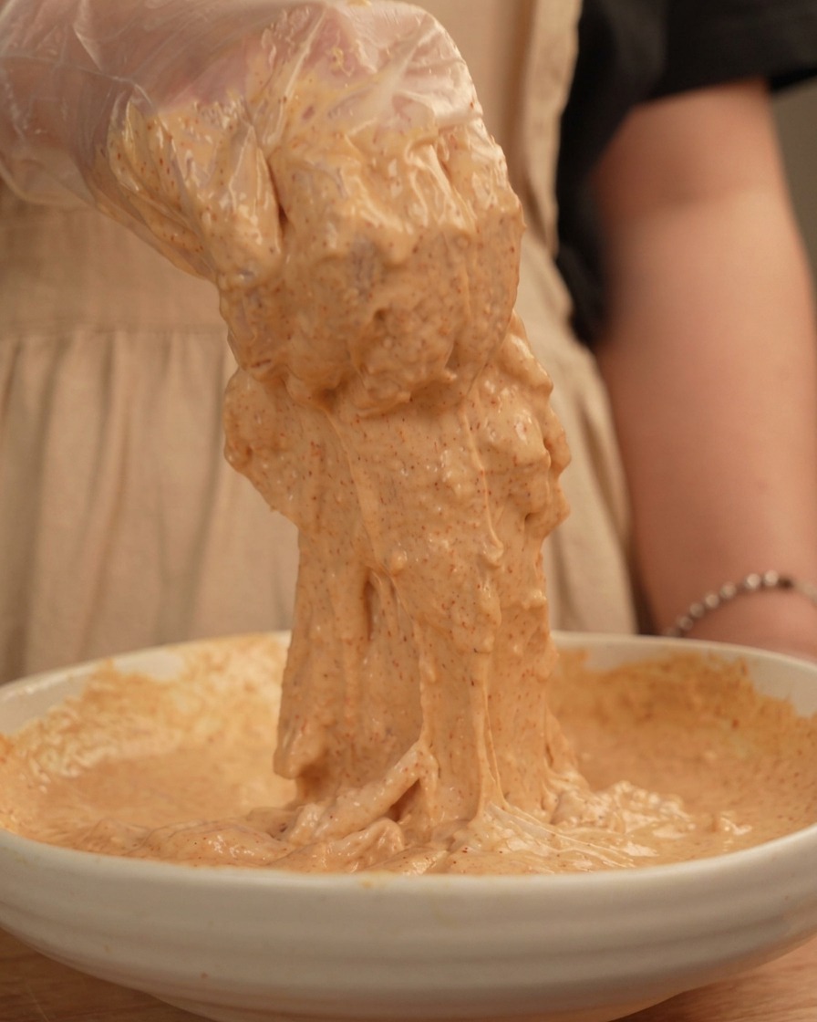 A gloved hand lifting a piece of raw chicken heavily coated in a thick, sticky, pale orange batter.