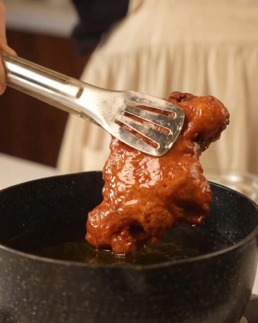 Metal tongs lifting a large, extra-crispy, golden-brown piece of fried chicken out of a pan of hot oil.