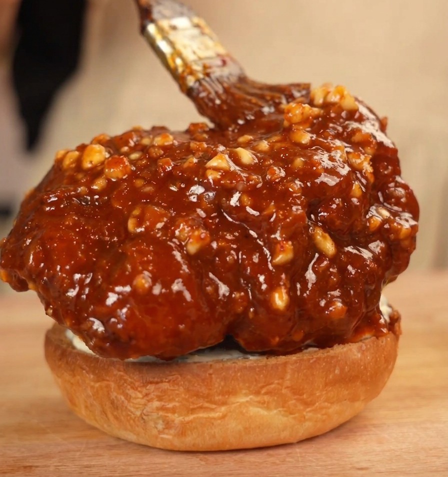 A large, sauce-coated fried chicken patty sitting on a bottom burger bun, being brushed with extra peanut chili sauce.