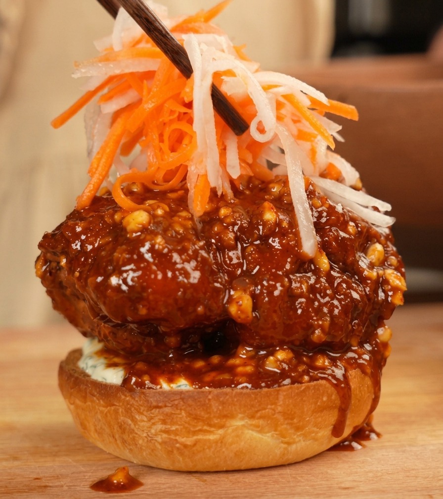 Chopsticks placing a colorful pile of julienned white radish and carrots on top of a saucy fried chicken burger patty.