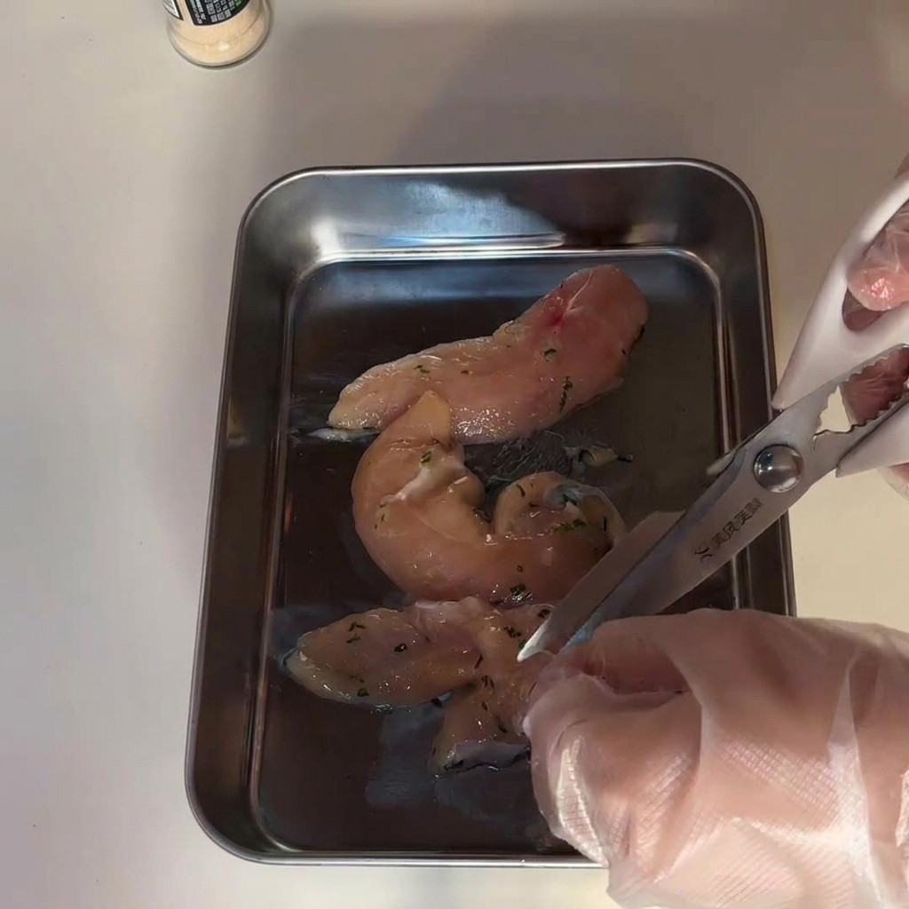 Hands wearing clear plastic gloves using kitchen scissors to cut raw chicken breast pieces on a stainless steel tray.