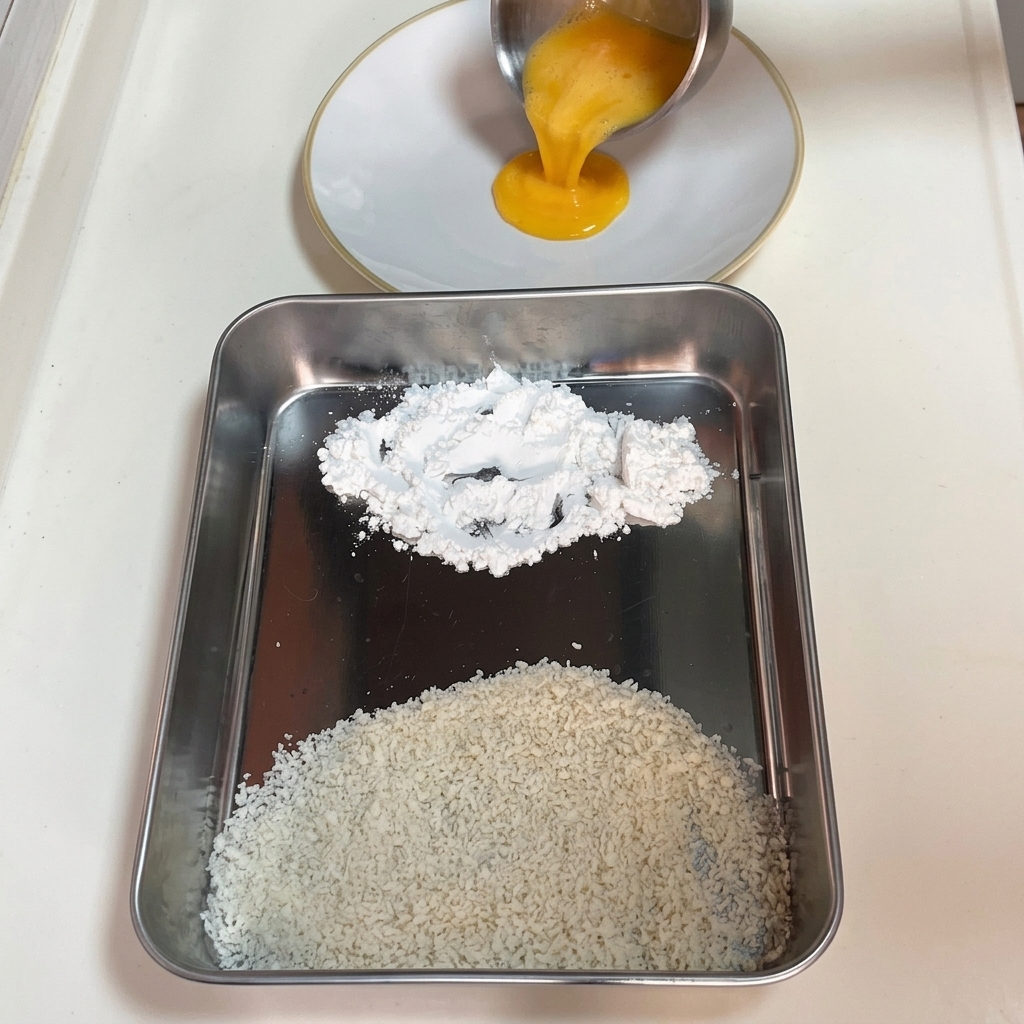 Pouring beaten yellow egg into a white plate, placed next to a metal tray holding separate mounds of white cornstarch and panko breadcrumbs.